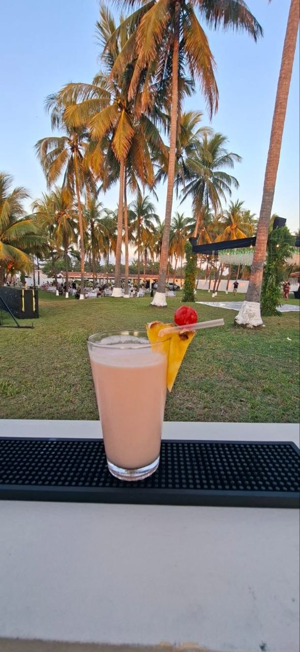 Cóctel decorado con piña y cereza sobre la barra, con palmeras al atardecer de fondo