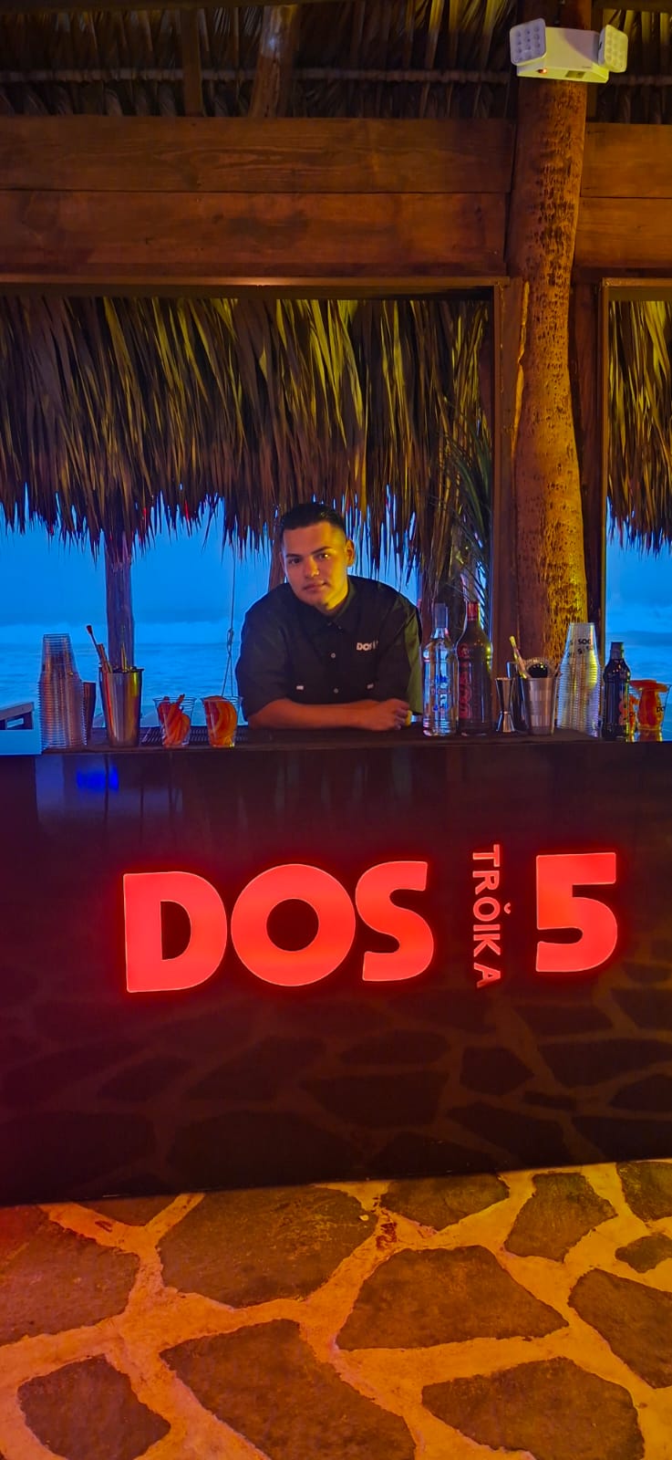 Carlos Ramírez detrás de una barra iluminada con el letrero «Dos 5 Tröika», junto al mar al anochecer